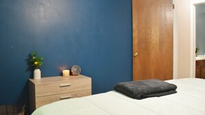 Ein Schlafzimmer mit einem Bett, einem Nachttisch aus Holz, einer weißen Vase mit Pflanze, einer angezündeten Kerze und einer Uhr.