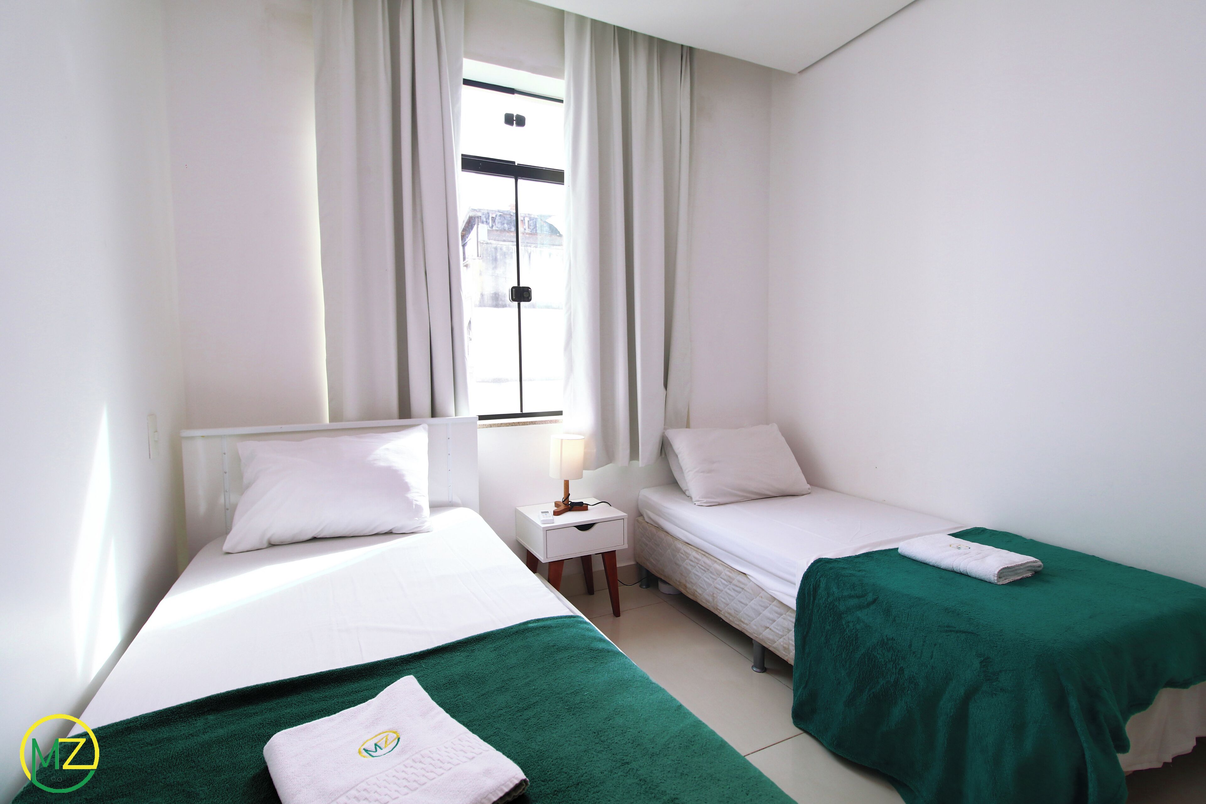 A hotel room with two beds, each with white bedding and green throw blankets. There is a small bedside table with a lamp and a window with white curtains.