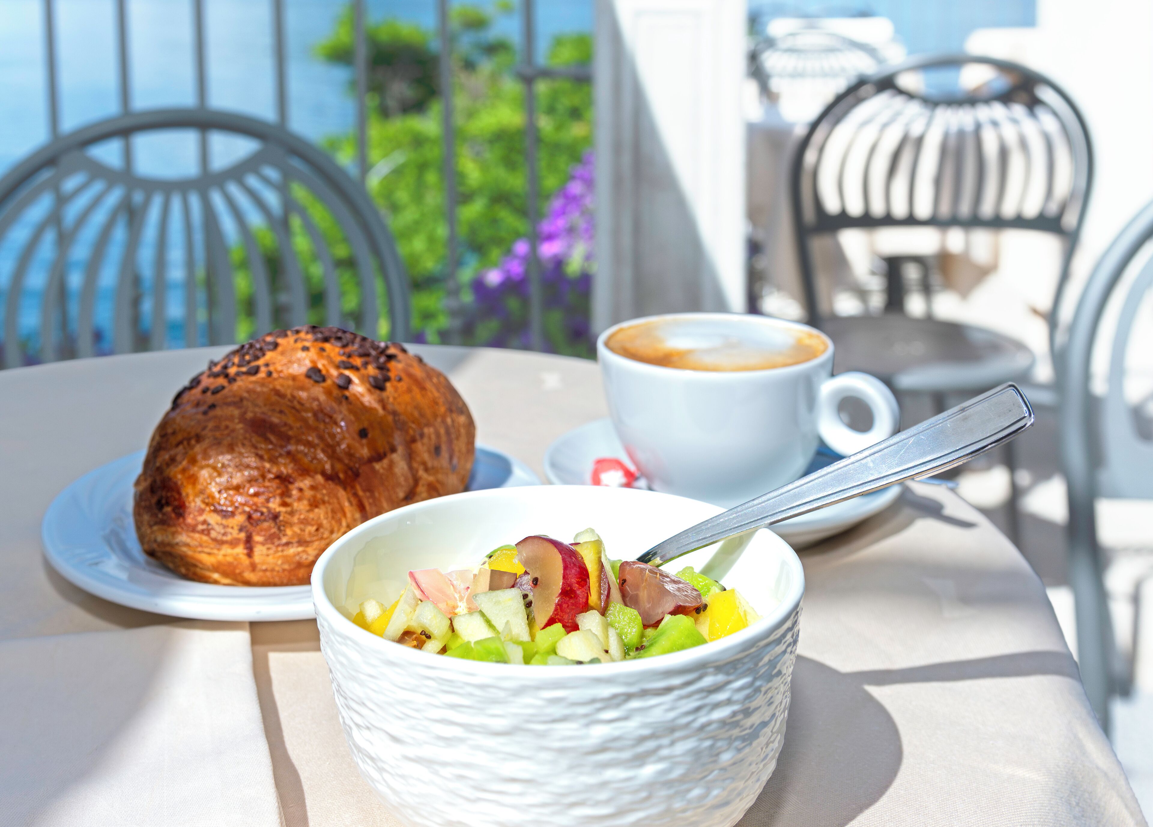 Ein Tisch mit einem Croissant, einer Obstsalatschale und einer Kaffeetasse auf einer Terrasse mit Metallstühlen und Blick ins Grüne.