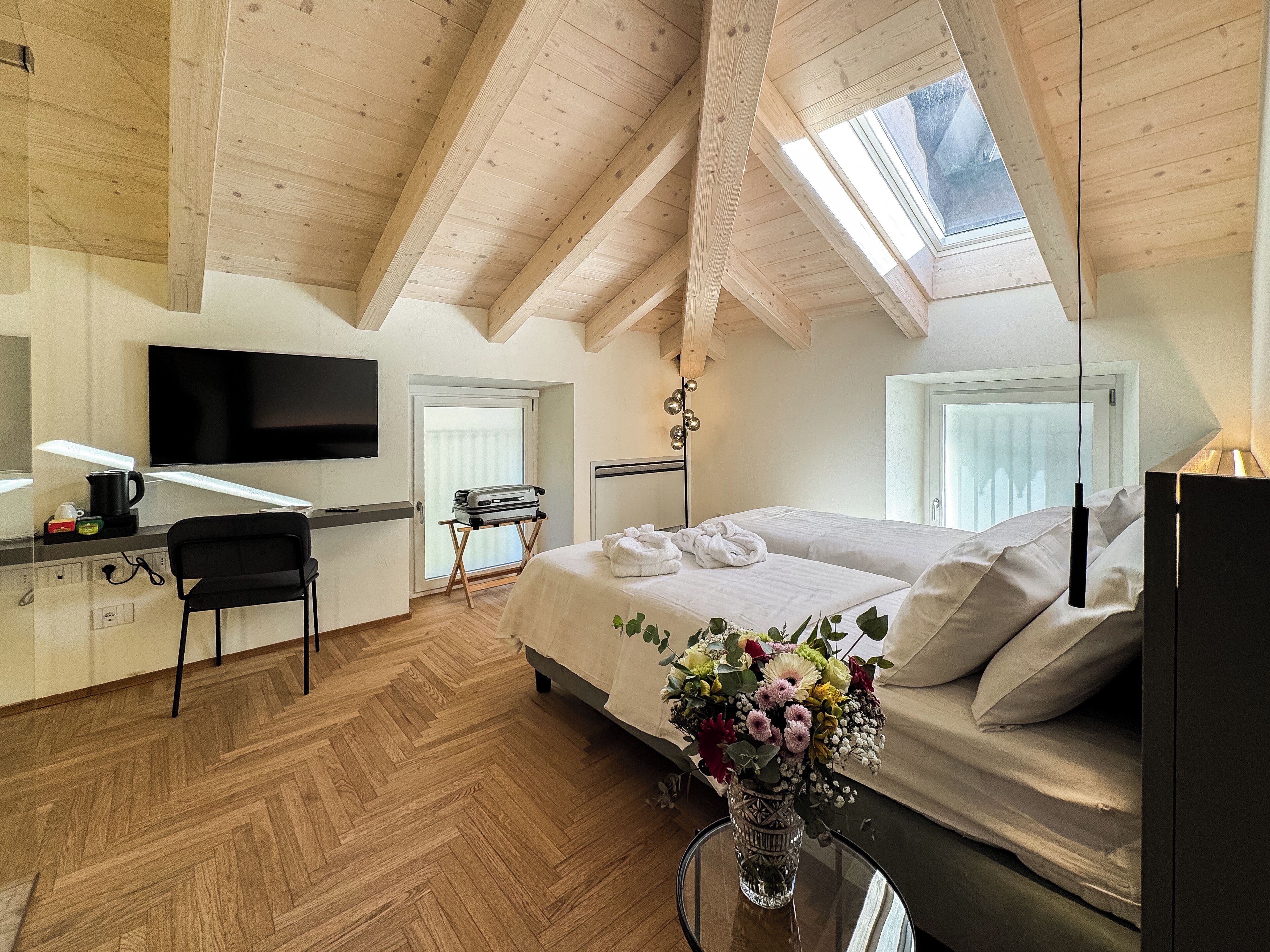 A modern hotel room with a wooden ceiling, a bed with white linens, a desk with a flat-screen TV, a chair, and a vase with flowers.