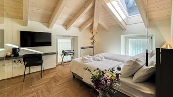 A modern hotel room with a wooden ceiling, a bed with white linens, a desk with a flat-screen TV, a chair, and a vase with flowers.
