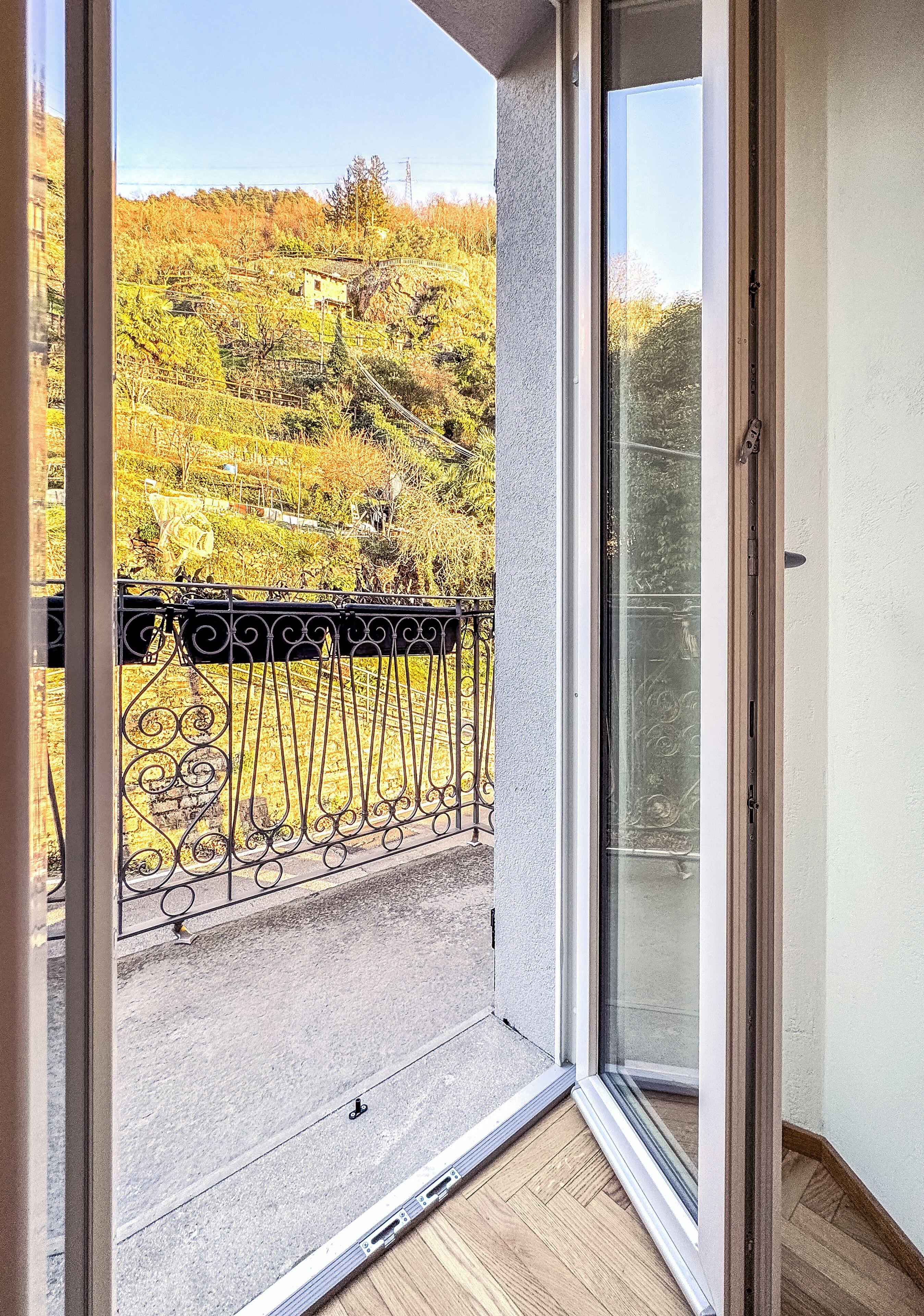 Un balcon doté d’une rambarde en métal, d’une porte vitrée et offrant une vue sur un paysage vallonné, avec des arbres et un bâtiment.