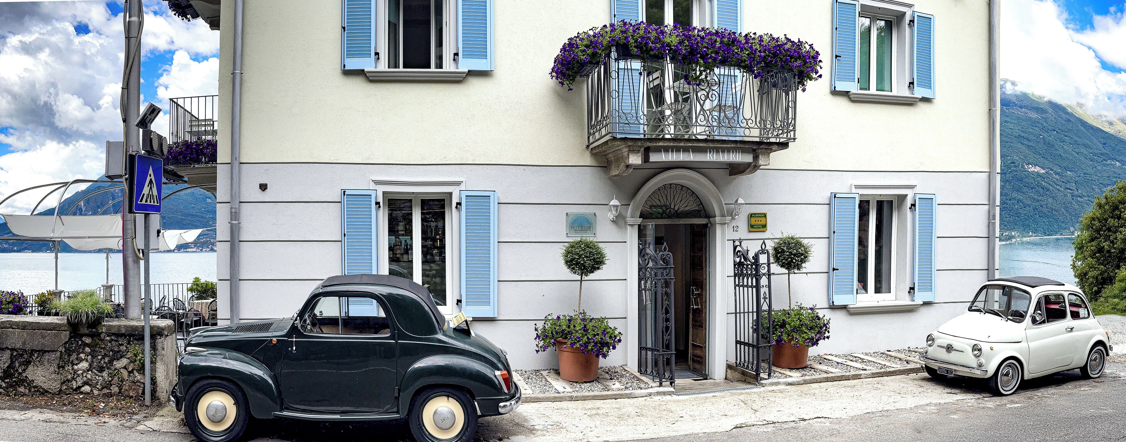 Ein Hotel mit Balkon, lila Blumen, einem geparkten Oldtimer davor und Bergblick im Hintergrund.