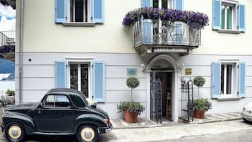 Ein Hotel mit Balkon, lila Blumen, einem geparkten Oldtimer davor und Bergblick im Hintergrund.
