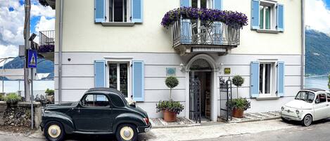 Ein Hotel mit Balkon, lila Blumen, einem geparkten Oldtimer davor und Bergblick im Hintergrund.
