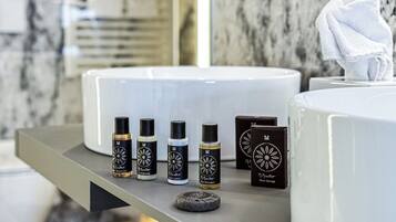 Et badeværelse med en hvid vask, et spejl og diverse toiletartikler, herunder brusergel, conditioner, bodylotion, hårshampoo og en skobørste.