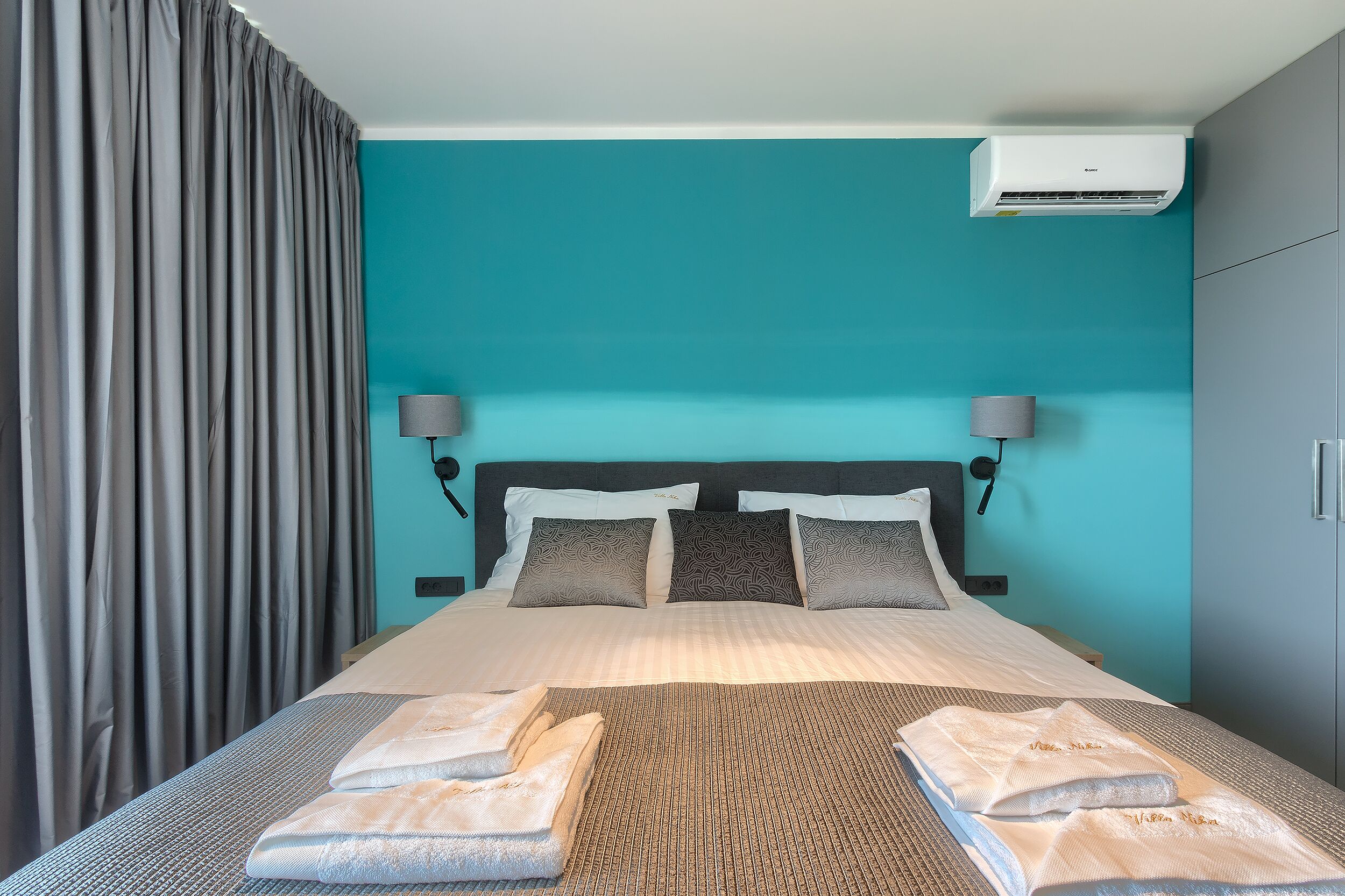 A hotel room with a bed, two wall-mounted lamps, a ceiling air conditioner, and a teal wall.
