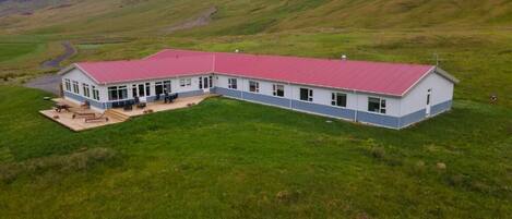 A long, single-story building with a red roof, situated on a grassy hill with mountains in the background.