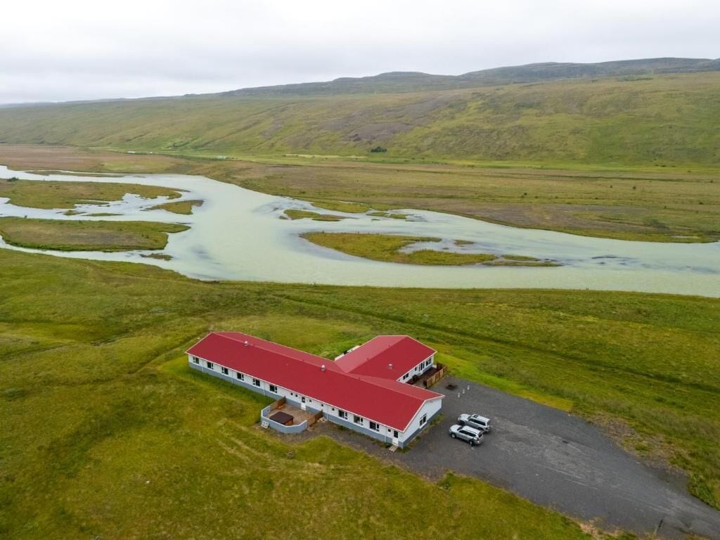 A two-story building with a red roof, surrounded by green fields and a river.