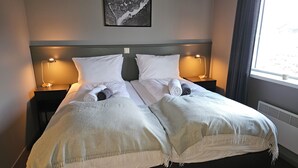 A double bed with white linens, two bedside tables with lamps, a framed picture on the wall, and a window with curtains. - Long Valley Lodge  (Blönduós)