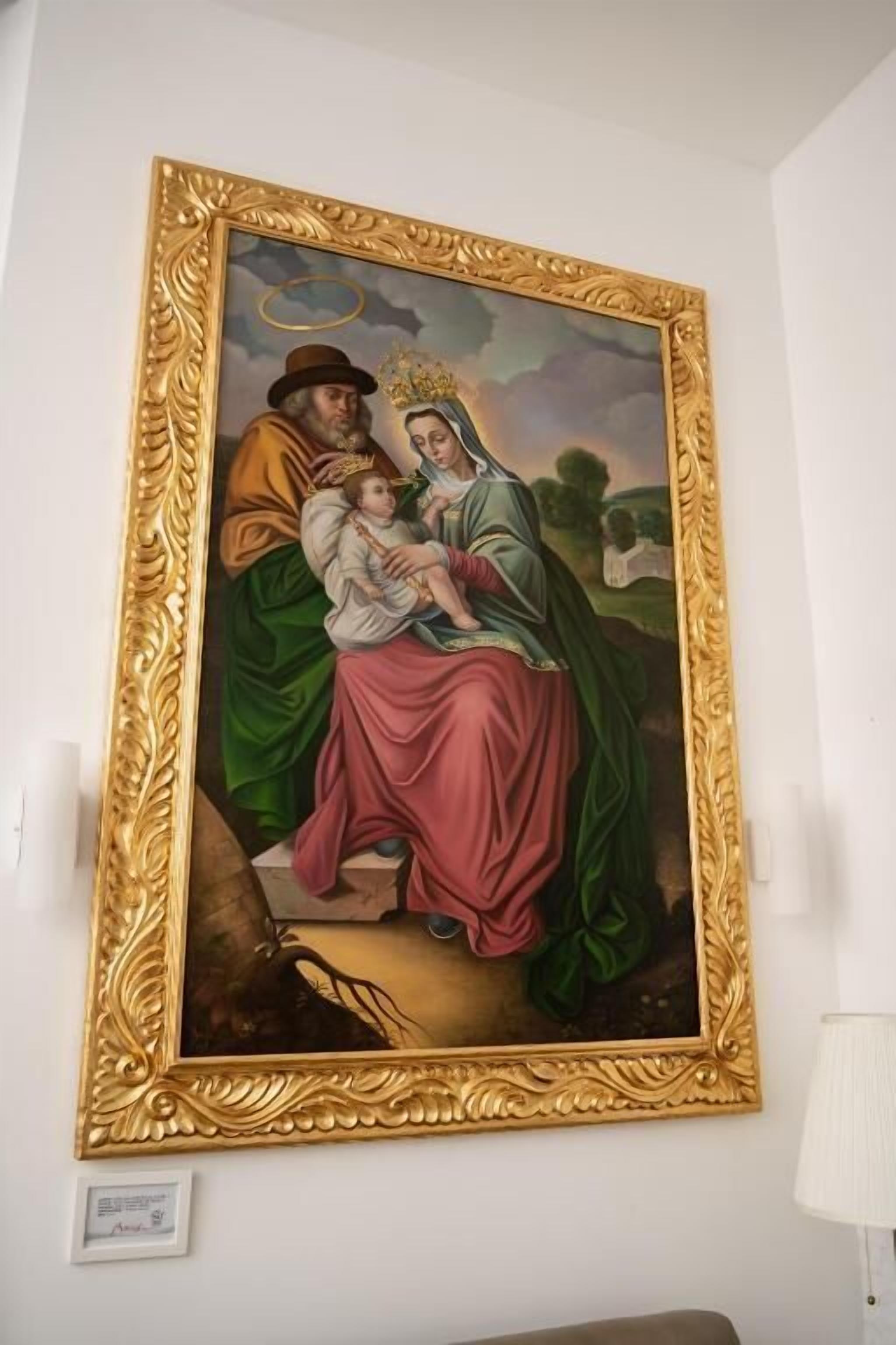 A framed painting of a religious scene with a halo, a child, and a woman in a green robe, mounted on a white wall.