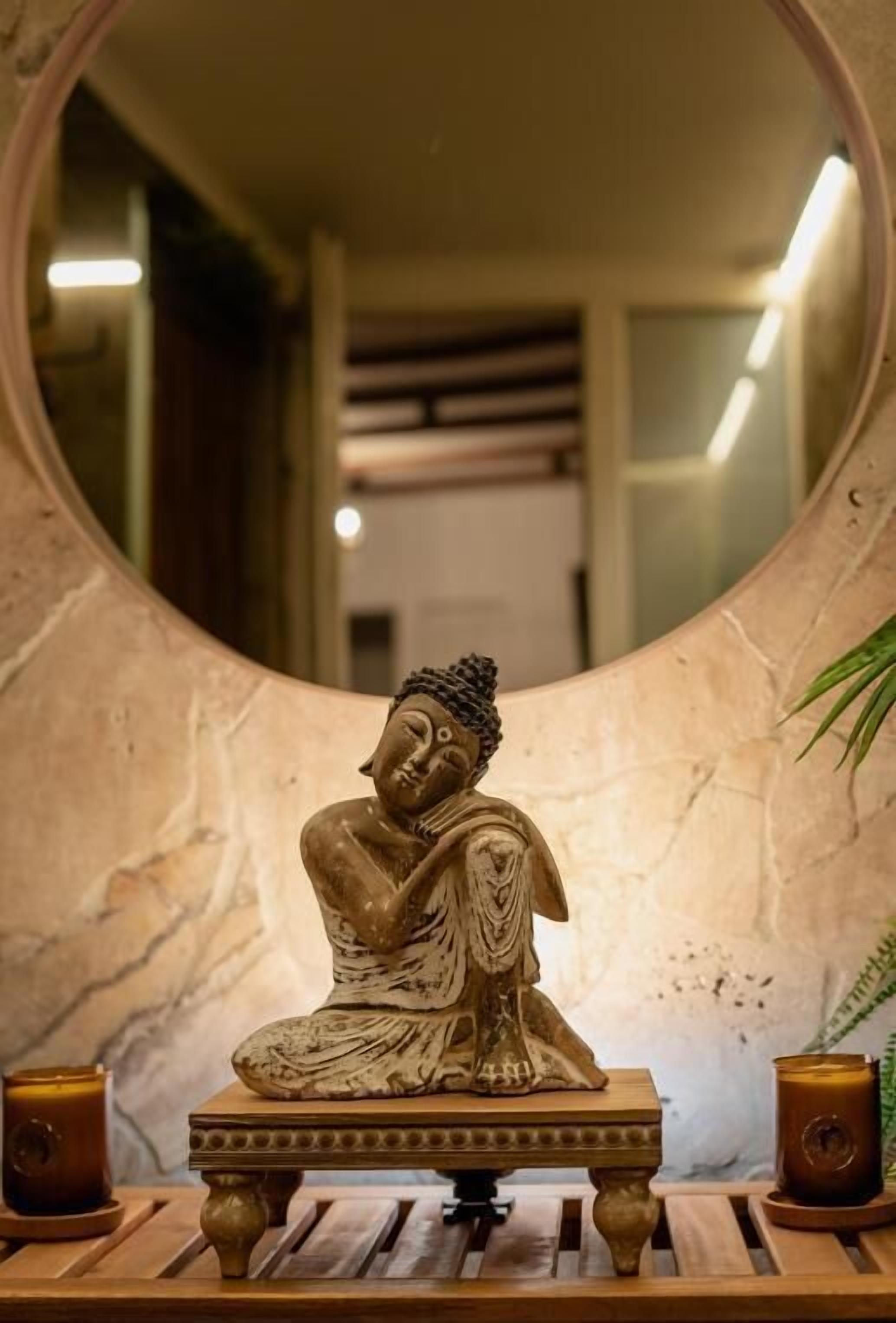 A Buddha statue on a wooden platform with two candles, set against a marble wall and a mirror.
