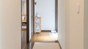 Un estrecho pasillo con suelo de madera y paredes blancas. Hay una puerta a la izquierda y una estantería con objetos a la derecha.