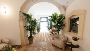 A spacious hallway with a checkered floor, two large potted plants, a beige armchair, a small round table, and a decorative vase. - Studio 1 apartment in Ortigia (Siracusa)