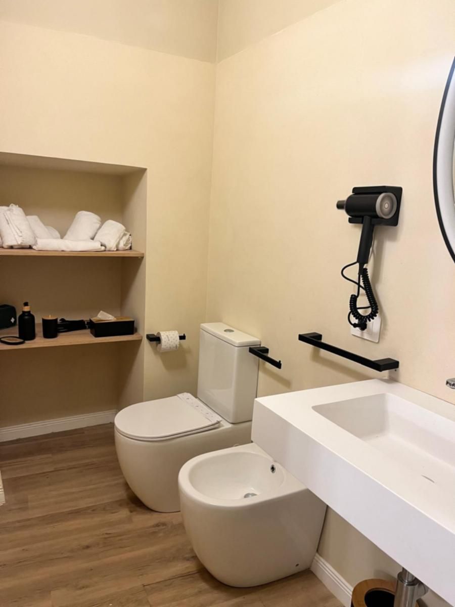 Baño pequeño con inodoro, bidé y lavamanos. Se observan toallas y artículos de aseo en una repisa.