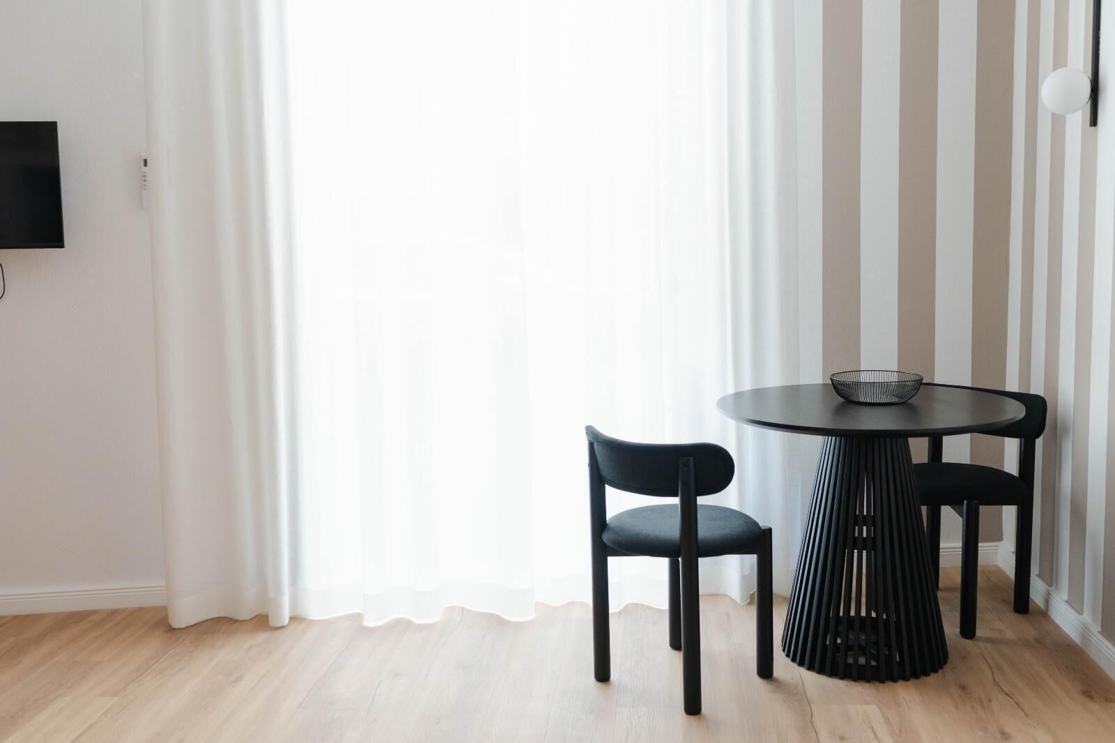A modern room with a round table and two chairs, a television on the wall, and a window with white curtains.