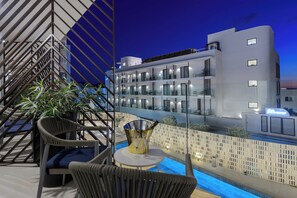 Un balcón moderno de hotel con mobiliario de mimbre, una piscina y vistas del exterior del edificio al anochecer.