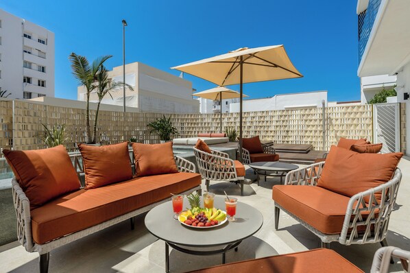 An outdoor seating area with wicker furniture and orange cushions, a round table with fruit and drinks, and a large umbrella providing shade. - Hotel Vibra Yamm Urban - Adults Only (Sant Antoni de Portmany)