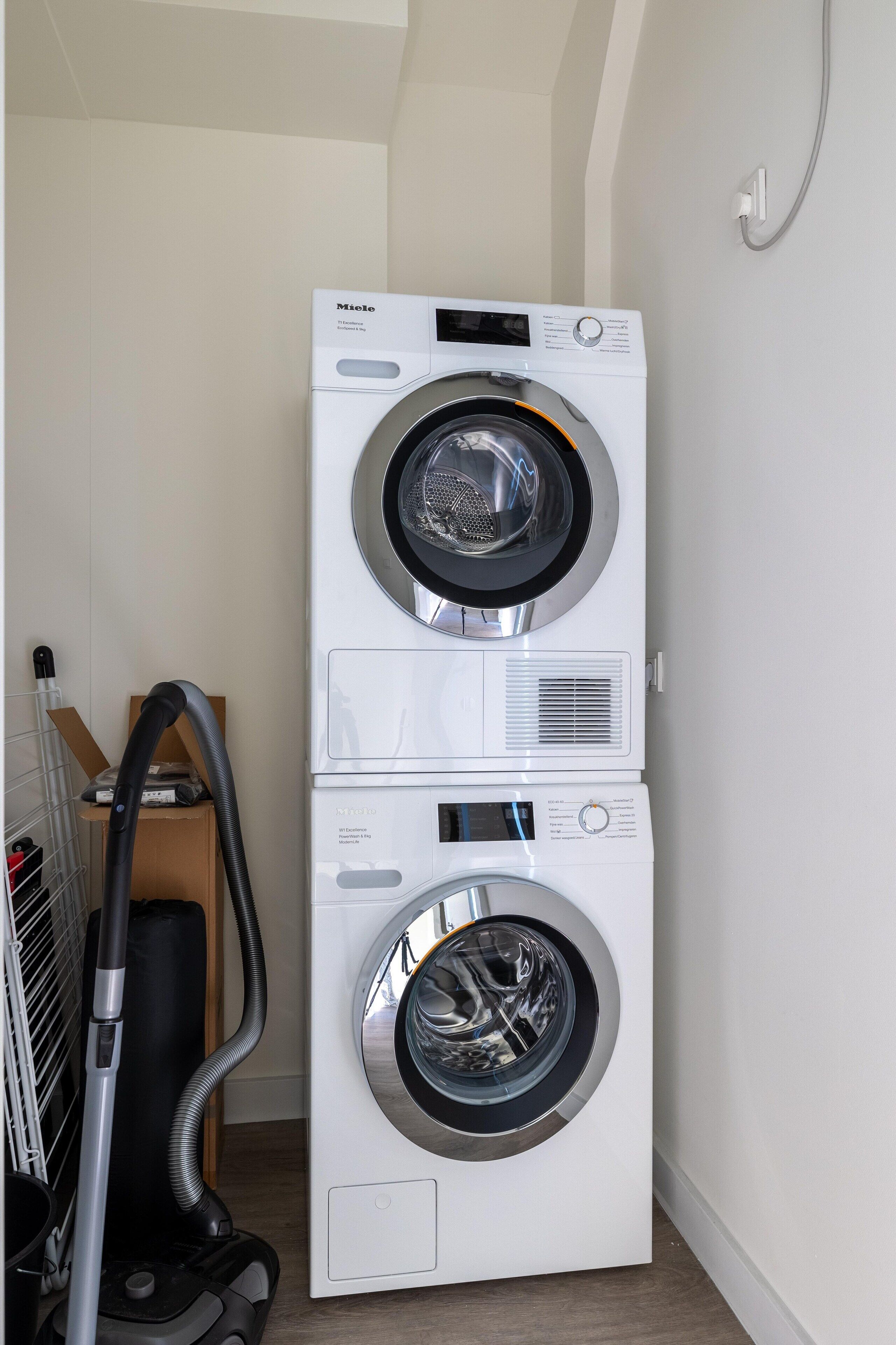 Une machine à laver et un sèche-linge superposés dans une buanderie, avec un aspirateur à proximité.