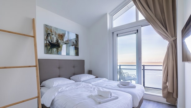 Ein modernes Hotelzimmer mit zwei Betten, einem großen Fenster mit Vorhängen, Blick aufs Meer und einem Bild an der Wand.