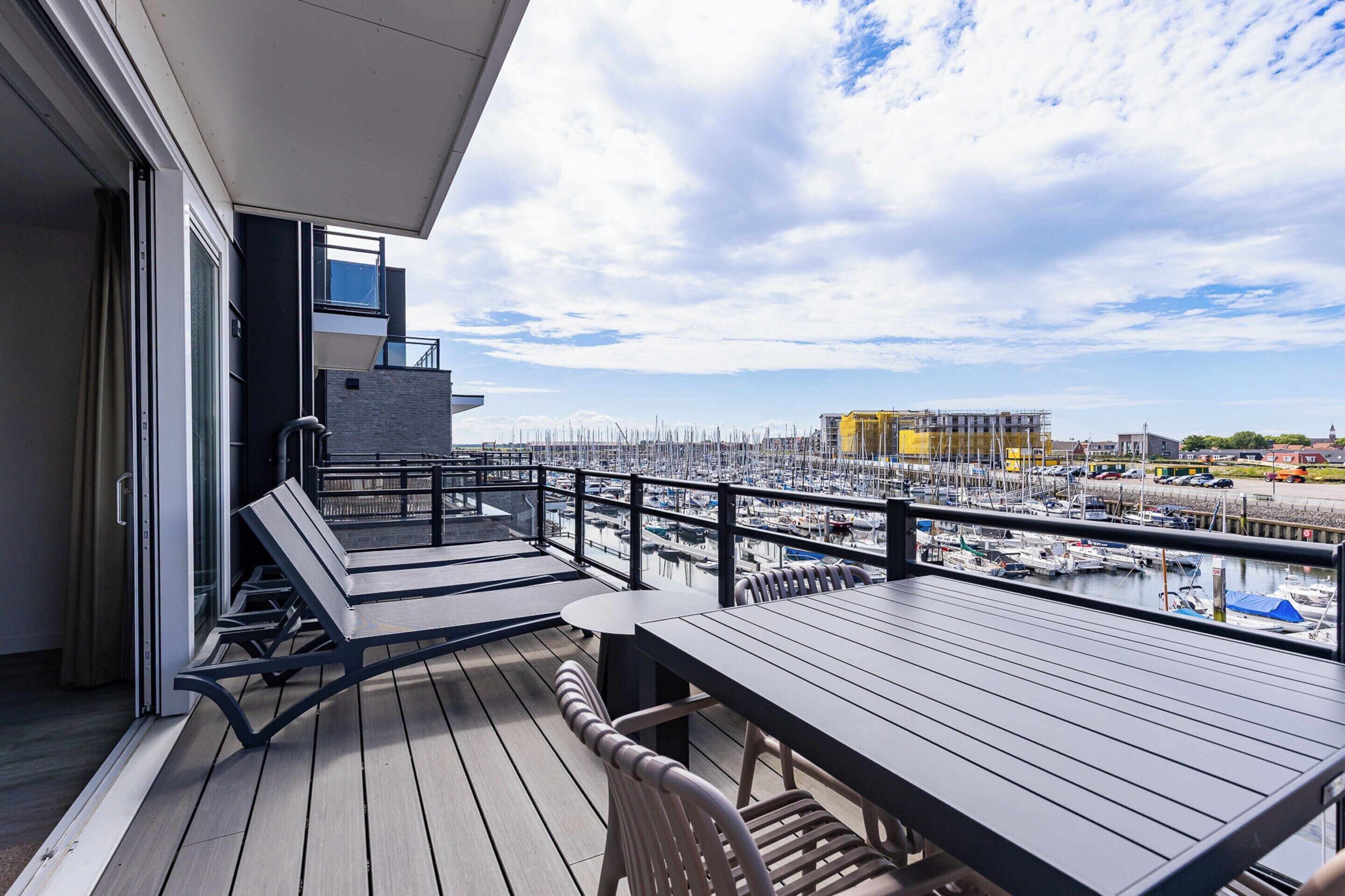 A balcony with a wooden deck, a table, and chairs, overlooking a marina with boats.