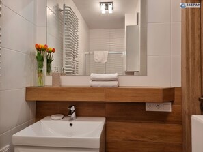 Ein modernes Badezimmer mit einem weißen Waschbecken, einer Holzanrichte und einem Spiegel darüber. Handtücher sind auf der Anrichte gestapelt und ein Blumenstrauß steht auf der Ablagefläche.