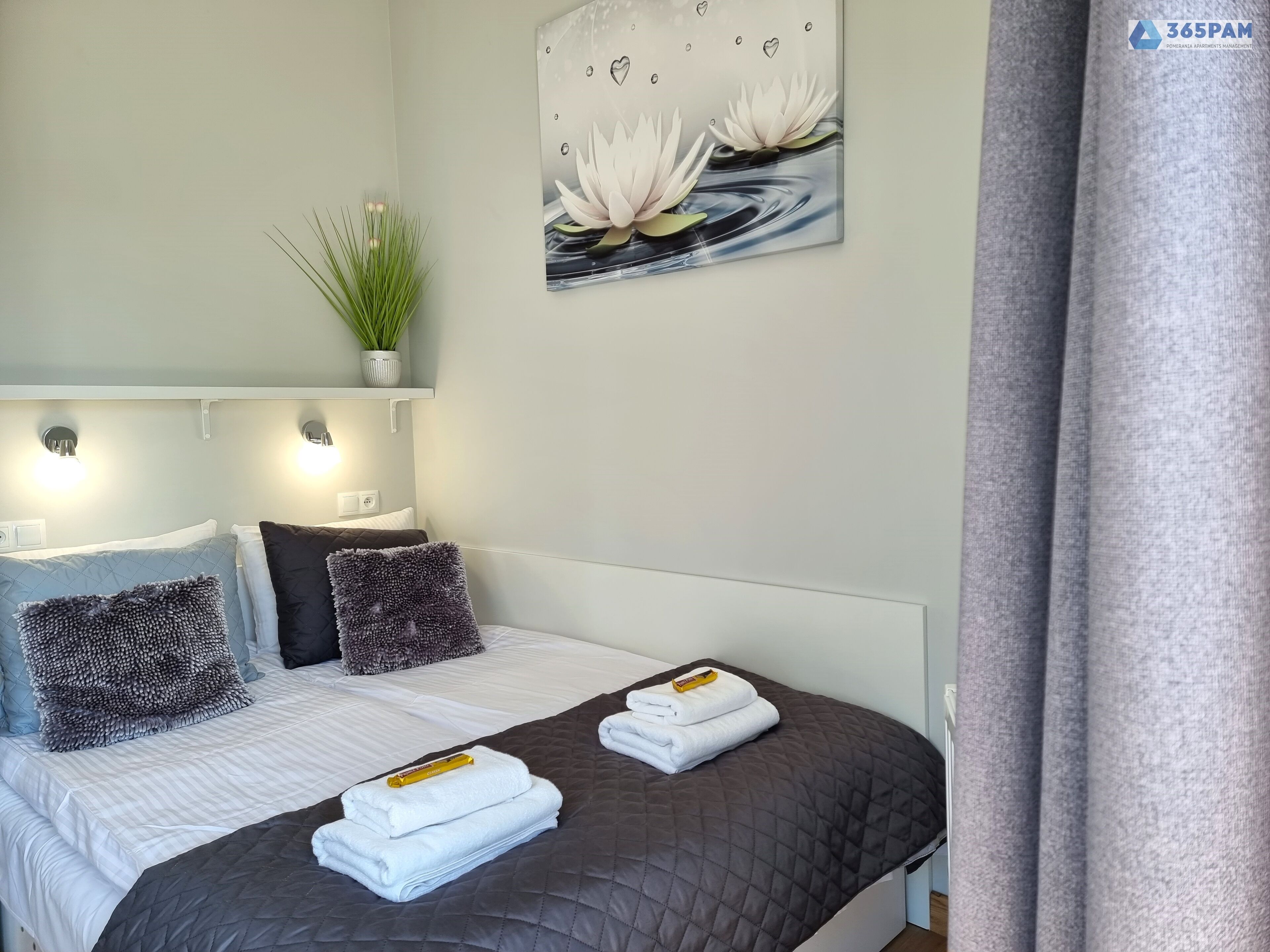 A neatly made bed with white and dark grey bedding, a decorative wall piece, and a potted plant.