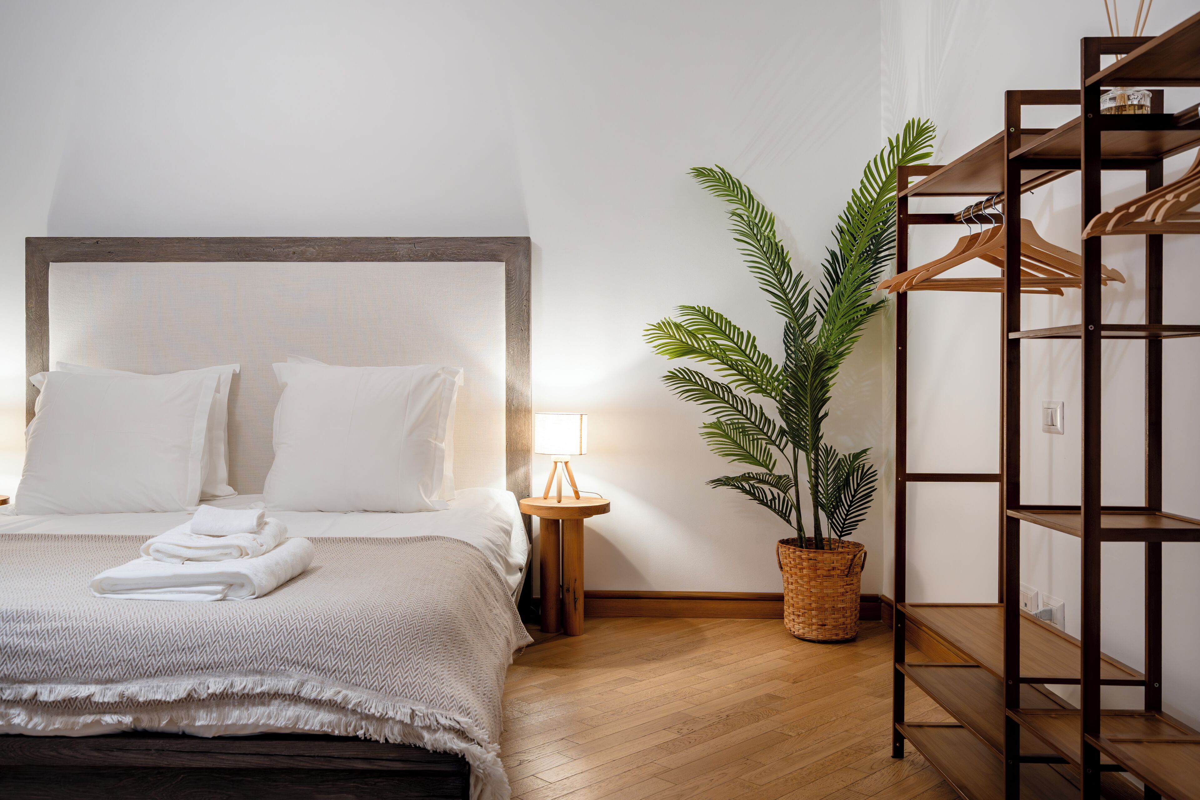Un letto rifatto con lenzuola bianche, una lampada da comodino, una pianta in vaso e una mensola in legno.