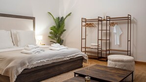 Un lit bien fait, recouvert d’une couverture blanche, une table de chevet en bois avec un vase, un petit pouf rond et un portant à vêtements en bois avec des cintres.