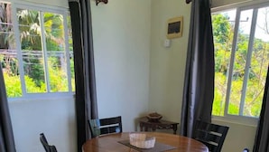 A round wooden dining table with six chairs, a small side table, and a window with curtains. - Sunset View Self Catering (Mahé Island)