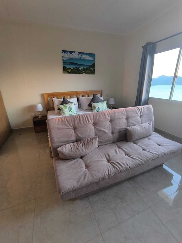 A modern hotel room with a large bed, a sofa, a painting on the wall, and a view through the window. - Sunset View Self Catering (Mahé Island)