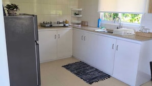 A compact kitchen with white cabinets, a gas stove, a refrigerator, and a window with a view of greenery. - Sunset View Self Catering (Mahé Island)