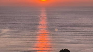 A sunset over a calm sea with silhouettes of trees and a house in the foreground.