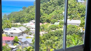 A room with a large window offering a view of a residential area, a body of water, and lush greenery. - Sunset View Self Catering (Mahé Island)