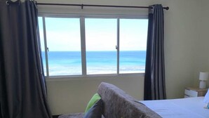 A room with a bed, a sofa with pillows, a window with curtains, and a view of the ocean. - Sunset View Self Catering (Mahé Island)