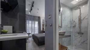 A modern bathroom with a glass shower enclosure, a vanity with a marble countertop, and a bed in the adjacent room.