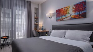 A hotel room with a large bed, a desk, a chair, a small table with a fruit bowl, and a painting on the wall.