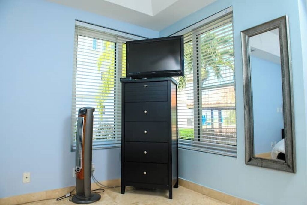 A room with a black dresser, a television on top, a standing heater, and a mirror on the wall.