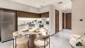 A modern kitchen with white cabinets, a stainless steel refrigerator, and a dining area with a round table and chairs. - Serene 2BR | Panoramic Sea View | Creek Edge | BMS (Dubai)