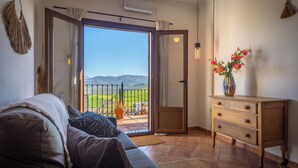 A living room with a sofa, a wooden side table with a vase of flowers, and a sliding glass door leading to a balcony with a view of green hills. - Boutique B&B Casa La Calera 15+/ Caminito del Rey-15 min/Ronda-25 min/Town-5 min (Teba)