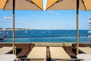 Zona junto a la piscina con sillones de descanso y sombrillas, con vista a un puerto deportivo lleno de veleros.