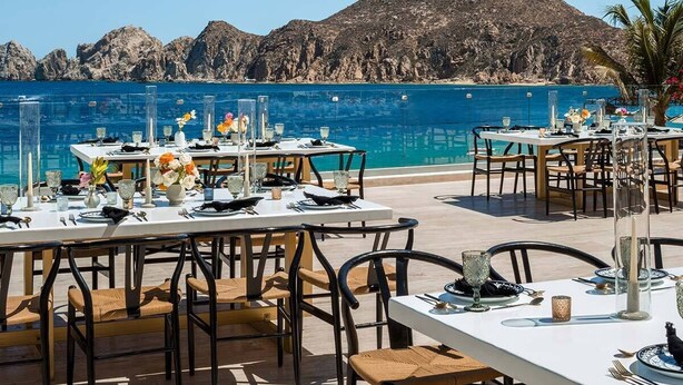 An outdoor dining area with tables set for two, overlooking a clear blue sea and rocky hills. - Mobility/Hearing Accessible One-Bedroom Deluxe (Cabo San Lucas)