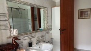 Un baño con tocador de mármol, lavamanos blanco, puerta de madera y un espejo, con una ducha a la vista en el fondo.