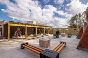 En udendørs patio med overdækket siddeplads, træbænke og krukker med planter.