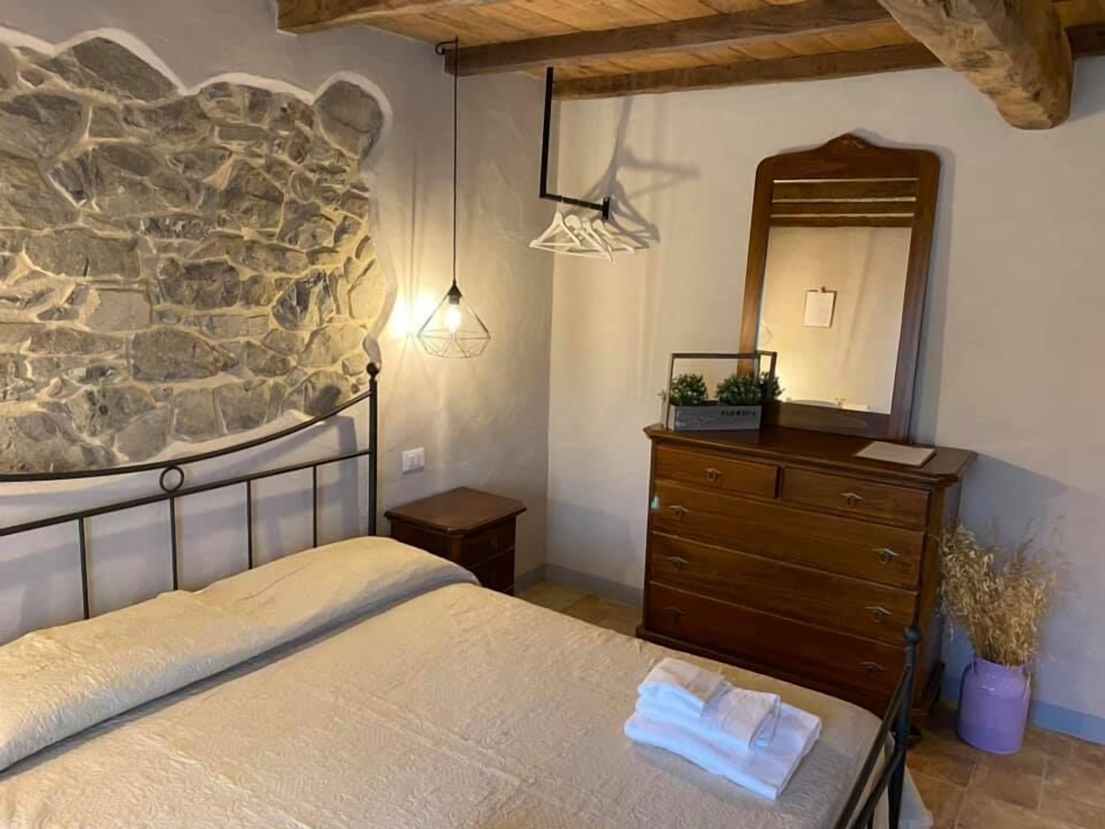 A bedroom with a stone wall, a bed with a white bedspread, a wooden dresser, and a hanging light fixture.