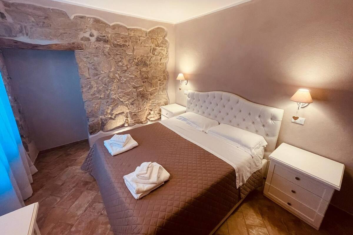Un dormitorio con pared de piedra, cama con cubrecama acolchado marrón, dos almohadas blancas y una mesita de noche con lámpara.
