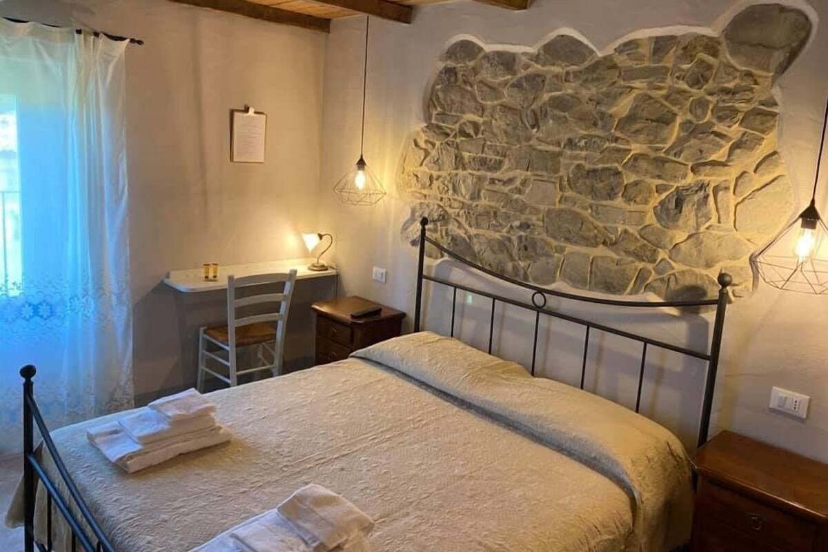 Un dormitorio con pared de piedra, una cama con cubrecama blanco, un escritorio con silla y dos lámparas colgantes.
