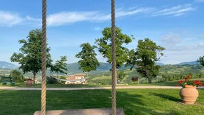 A wooden swing set in a grassy area with a view of a house and trees in the background. - L'OCA GIULIVA RELAIS  (Gaggio Montano)