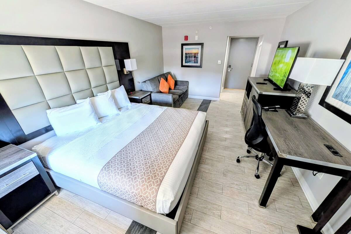A hotel room with a large bed, a desk with a computer, a TV, and a small seating area with a sofa and pillows.