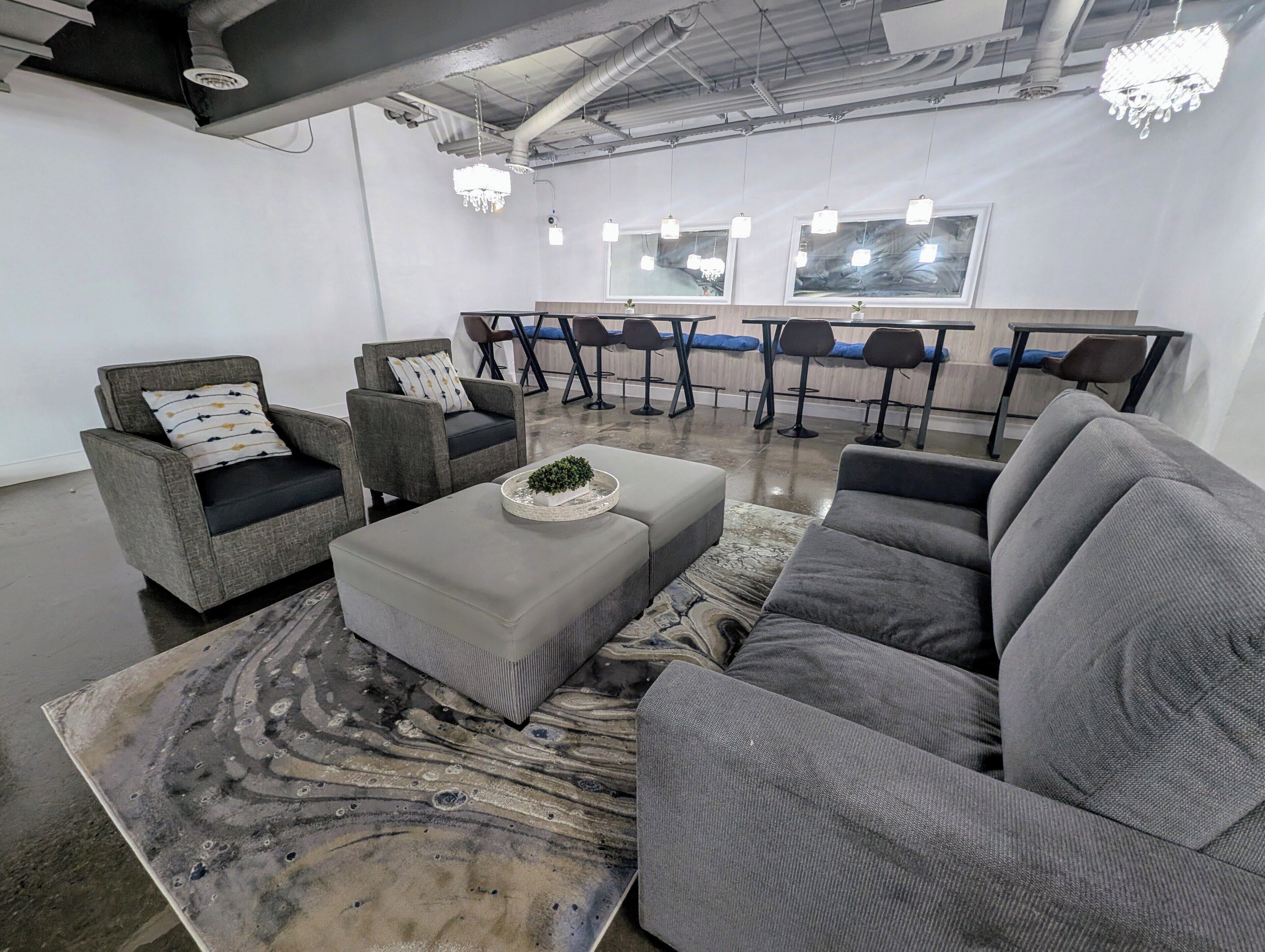 Un intérieur moderne avec des canapés gris, des fauteuils et une table basse. On aperçoit des tabourets de bar et une table à manger au fond.